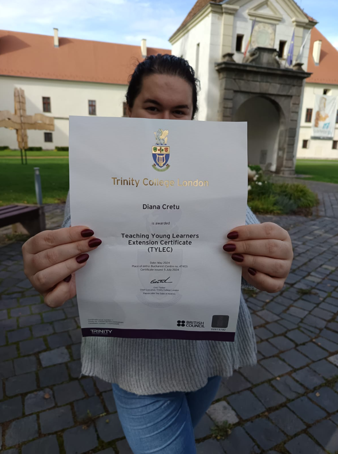 A woman holding a Teaching Young Learners Extension Certificate from Trinity College London, set against a background of a building and greenery.